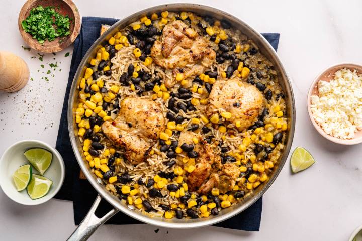 A skillet with seasoned chicken thighs, corn, black beans, and rice. Nearby, bowls of chopped herbs, lime wedges, and crumbled cheese enhance the dish.