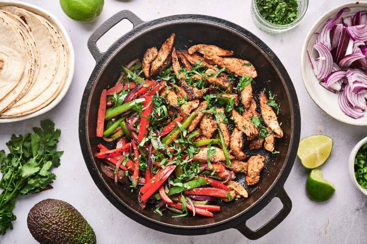 Pork fajitas with thin strips of pork, bell peppers, onion, cilantro, and limes in a cast iron skillet.
