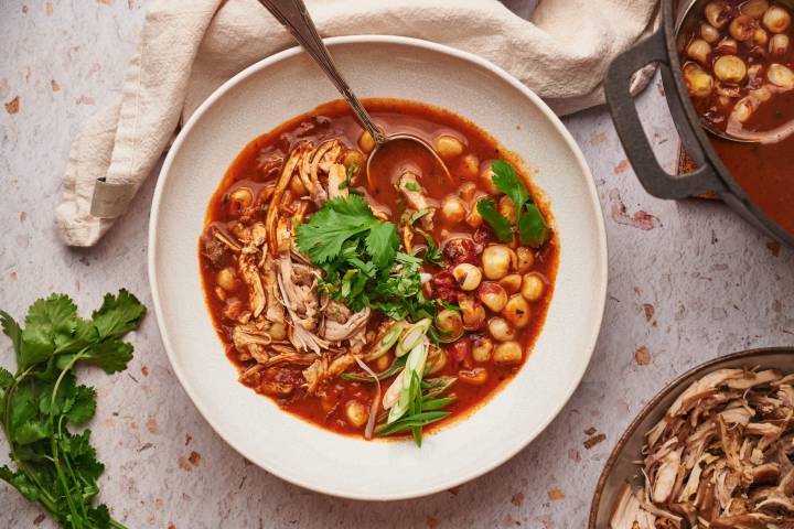 Red chicken pozole in a bowl with shredded chicken, hominy, red chile broth, avocado, and limes. 