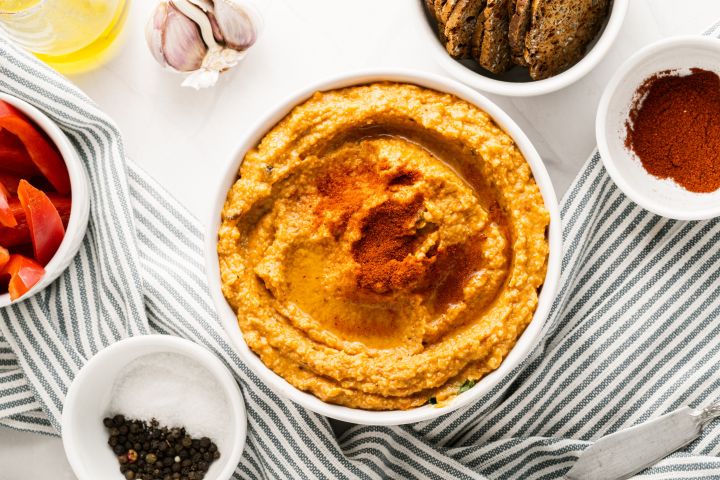 Roasted red pepper dip in a bowl drizzled with olive oil with garlic, crackers, and red bell pepeprs on the side.