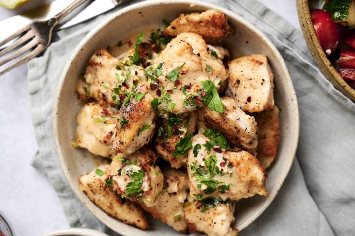 A bowl of golden-brown chicken pieces garnished with fresh parsley and red pepper flakes, set on a grey cloth next to a fork