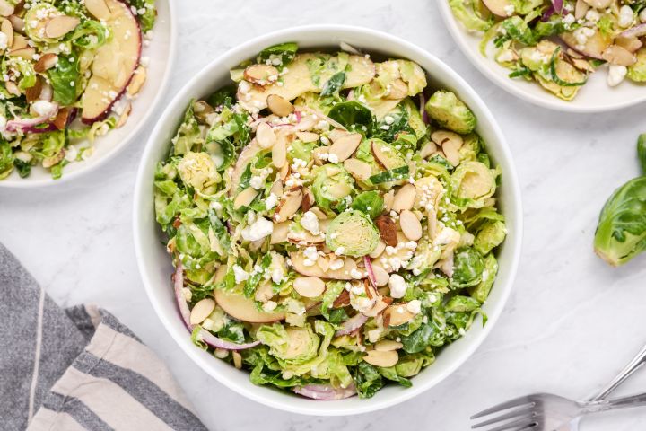 Shaved Brussels sprouts salad with creamy honey dijon dressing, sliced apples, goat cheese, almonds, and red onions in a bowl.