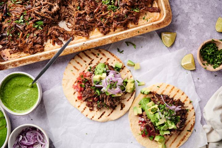 Crockpot carne asada served on tortillas with avocado, pico de gallo, red onion, and lime wedges.
