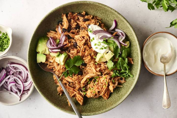 Chicken burrito bowl made with shredded chicken, rice, and black beans, topped with avocado, sliced red onion, sour cream, fresh cilantro, and chives. Small bowls of sour cream and red onion are arranged alongside the dish.