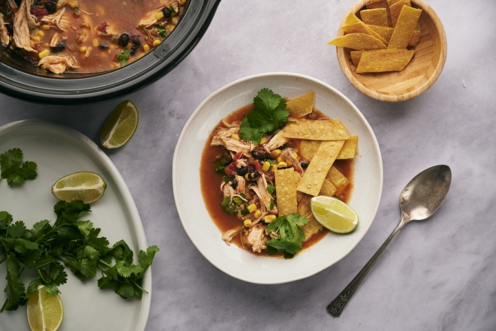 Slow cooker chicken tortilla soup with shredded chicken, beans, corn, cilantro, crispy tortilla strips, and lime wedges.