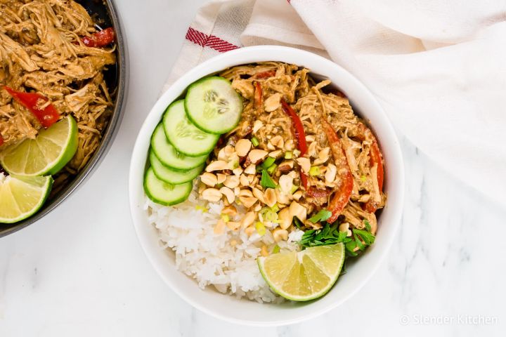 Slow Cooker Thai Peanut Chicken in a bowl with rice, cucumbers, chopped peanuts, and lime. 