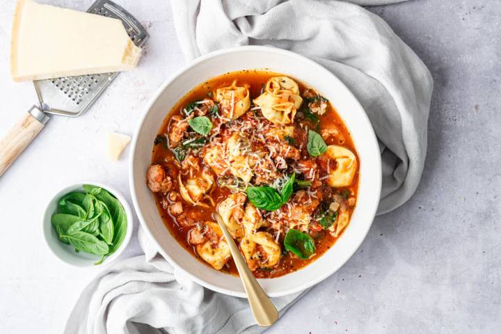 A warm and hearty bowl of Slow Cooker Tortellini Soup filled with tender tortellini, flavorful sausage, spinach, and tomatoes, topped with freshly grated Parmesan and basil for a comforting finish.