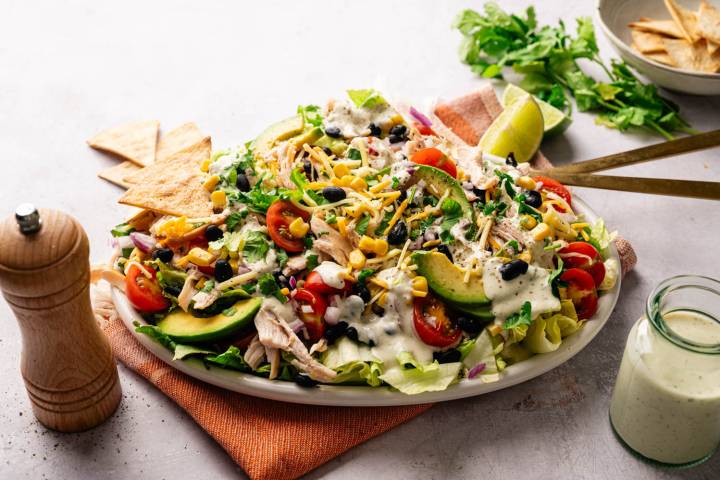 Angled view of Southwest chopped salad on an oval platter, garnished with lime wedges and cilantro, with a jar of dressing and tortilla chips beside it.