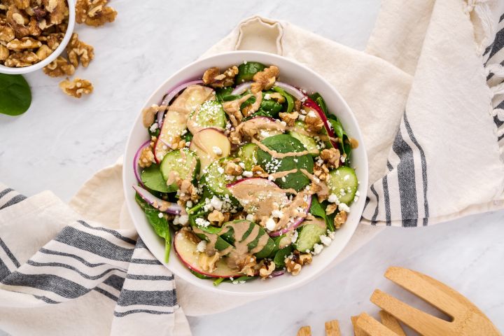 Spinach salad with creamy balsamic vinaigrette, goat cheese, apples, and walnuts in a white bowl with wooden salad tongs.