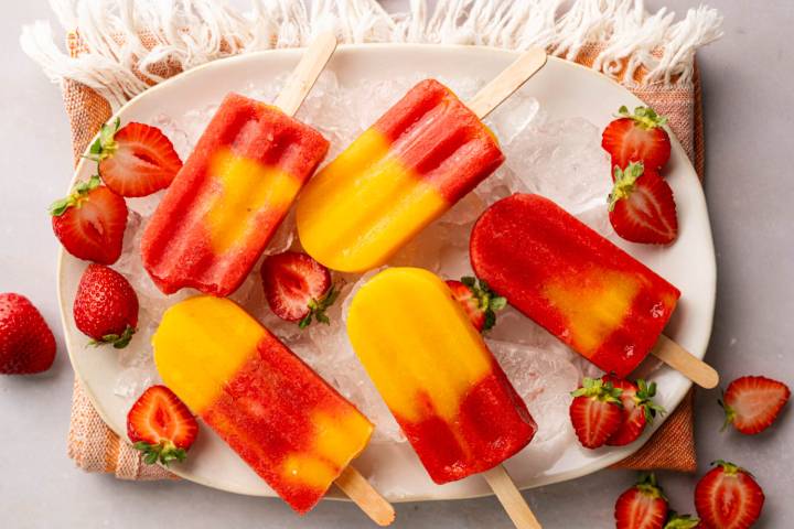 A platter of layered strawberry mango popsicles on crushed ice, garnished with fresh strawberries for a refreshing summer treat.