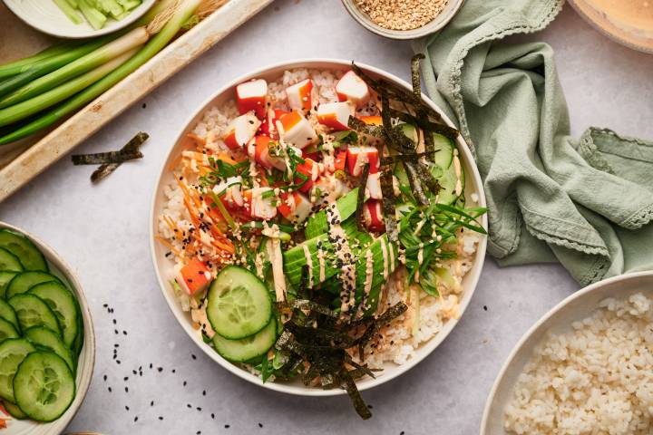 Sushi bowl with imitation crab, sushi rice, nori seaweed strips, cucumbers, carrots, and spicy mayo in a white bowl.