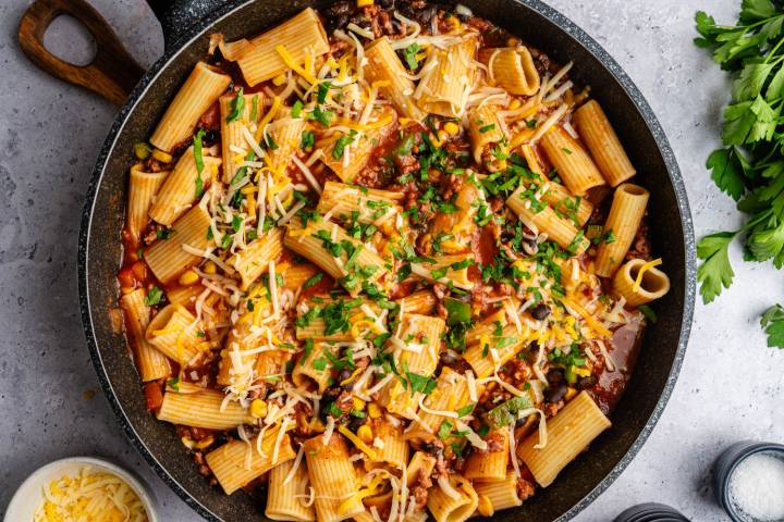 A pan of rigatoni pasta with tomato sauce, ground meat, corn, and black beans, topped with melted cheese and parsley, on a gray surface.