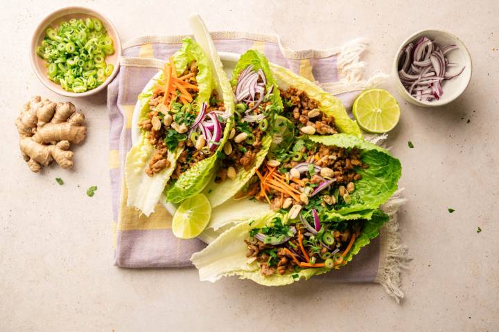Thai turkey lettuce wraps served in crisp romaine leaves and topped with shredded carrots, red onion, green onions, cilantro, peanuts, and lime wedges on a white plate.
