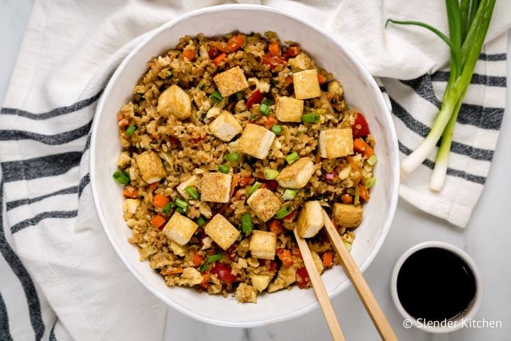 Tofu cauliflower fried rice with assorted vegetables, green onions, and soy sauce in a bowl with chopsticks.