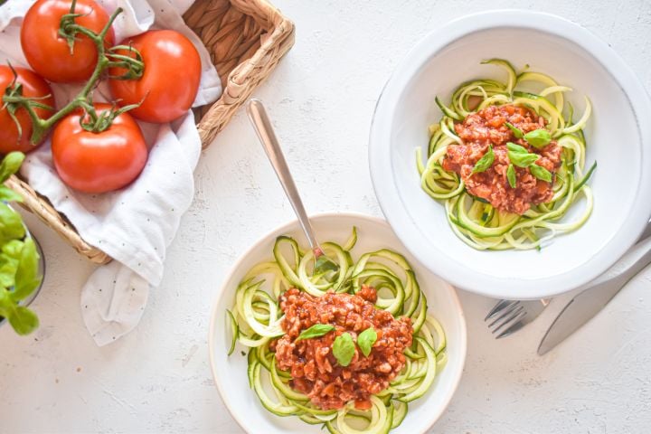 Zucchini noodle bolognese with zucchni noodles, turkey bolognese sauce, and basil on a plate with tomatoes on the side.