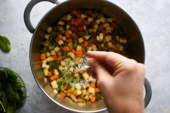 Zucchini and green beans being adding to a soup pot with salt and pepper.