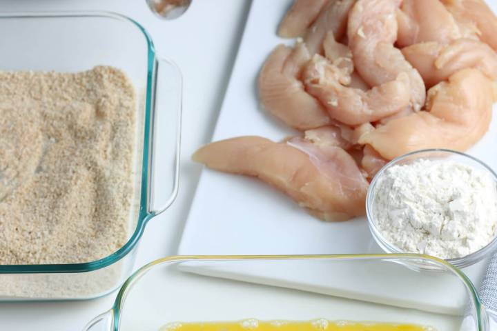 Chicken tender breading station with eggs, raw chicken, flour, and breadcrumbs.