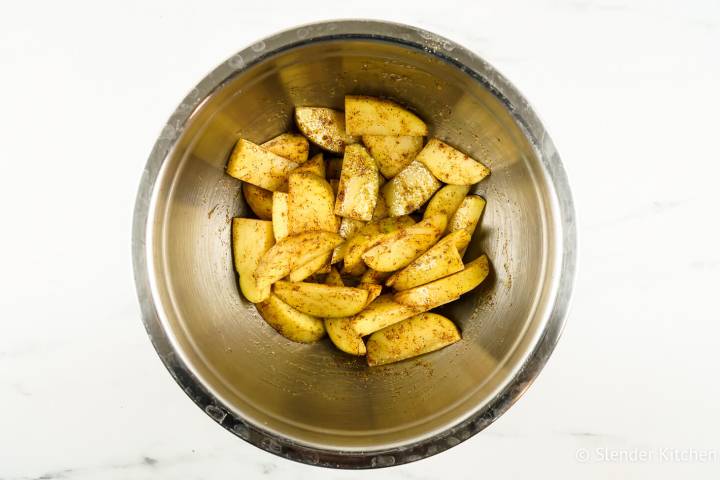 Potato wedges tossed with olive oil, paprika, garlic powder, salt, and pepper in a silver bowl.