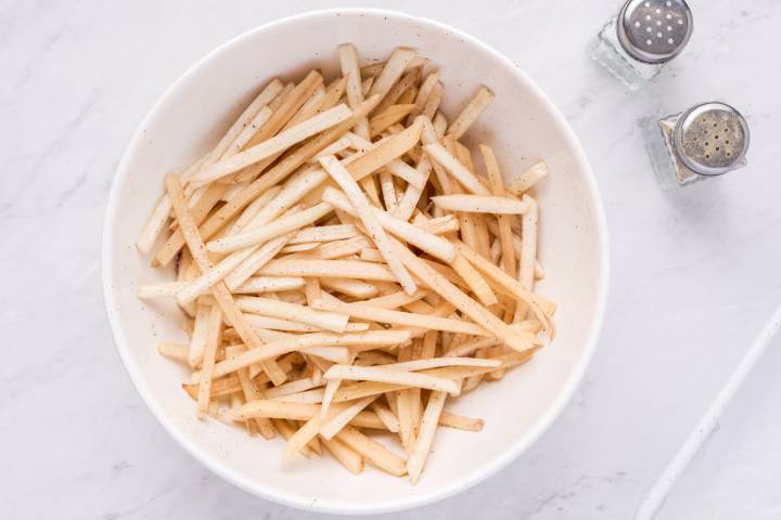 Potatoes and turnips cut into thin fries tossed with olive oil and seasoning in a white bowl.