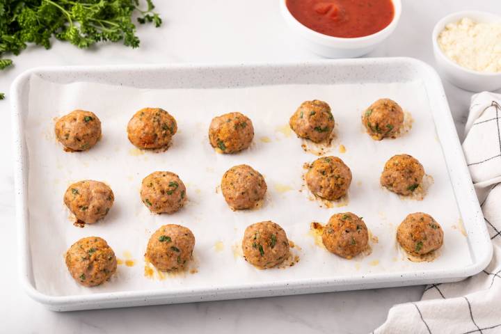Baked turkey meatballs on baking sheet with parsley and sauce on the side.