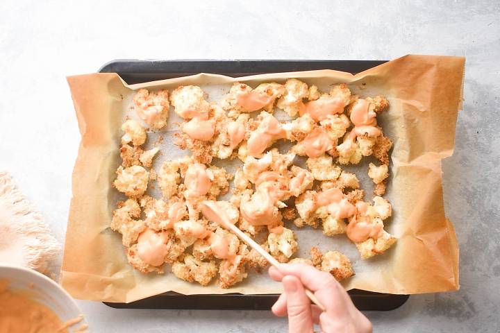 Baked breaded cauliflower being tossed with bang bang sauce on a baking sheet.