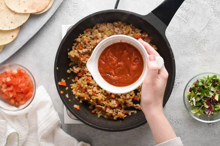 Red salsa being added to a skillet of ground taco beef.