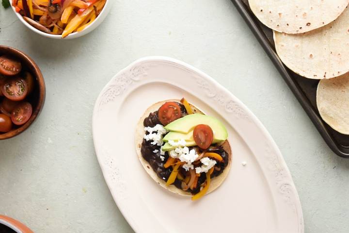Crispy corn tortilla topped with black beans, tomatoes, avocado, peppers, and onions on a white plate.