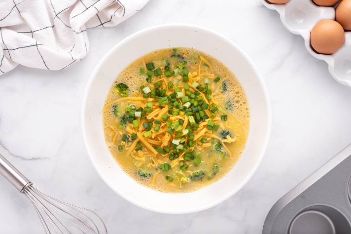 Eggs whisked in a bowl with broccoli, green onions, and cheddar cheese.