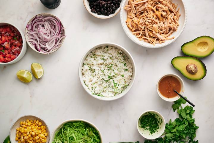Ingredients for carnitas burrito bowls including rice, carnitas, beans, corn, and toppings.