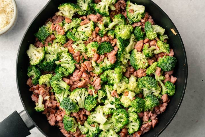 A skillet filled with vibrant green broccoli florets and browned ground sausage pieces. A bowl of grated cheese is partially visible on the side.