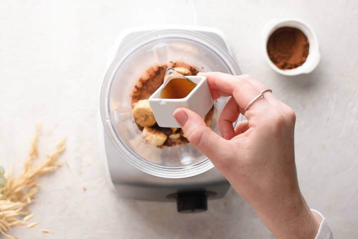 Maple syrup being added to a blender with bananas, tofu, almond milk, and coca powder.