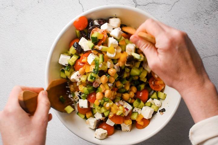 Chickpeas, chopped vegetables, herbs, feta, and olives being tossed together in white bowl.