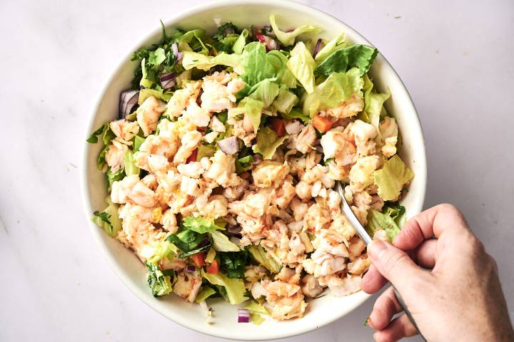 A close-up of a person's hand holding a spoon with chopped shrimp salad over a bowl.