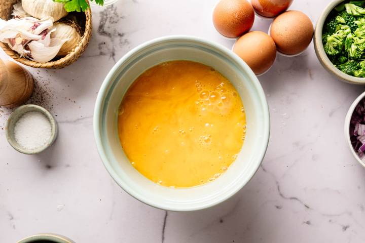 A bowl with beaten eggs on a marble counter surrounded by eggs, broccoli, garlic, salt, and pepper. Ingredients for a cooking preparation.