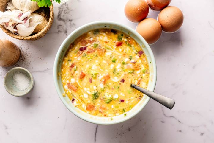 A bowl of colorful, mixed egg ingredients with tomatoes, onions, and herbs. Surrounded by whole eggs, garlic, and a salt dish on a marble countertop.