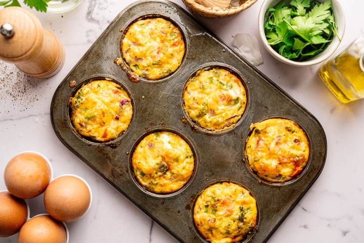 Muffin tin with six golden-brown egg muffins filled with veggies, on a marble counter. Nearby are eggs, parsley, a pepper grinder, and a bottle of oil.