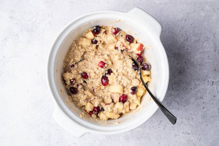A white bowl filled with oatmeal mixed with apple chunks and red cranberries, against a gray textured background. 