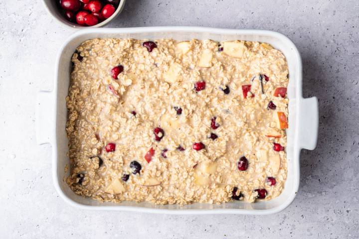 Baking dish with oatmeal mixture, mixed with cranberries and apple chunks, on a light countertop. A bowl of fresh cranberries is nearby.