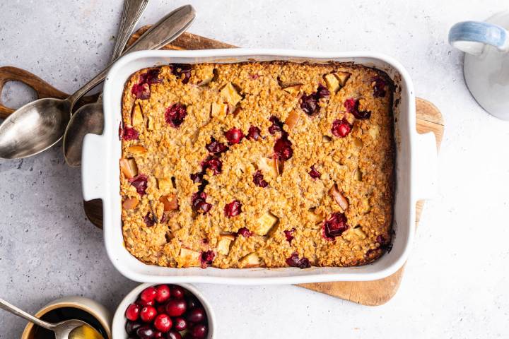 Baked oatmeal in a white dish, topped with cranberries and apple chunks. Surrounded by spoons and bowls of cranberries on a wooden board. Cozy and inviting.
