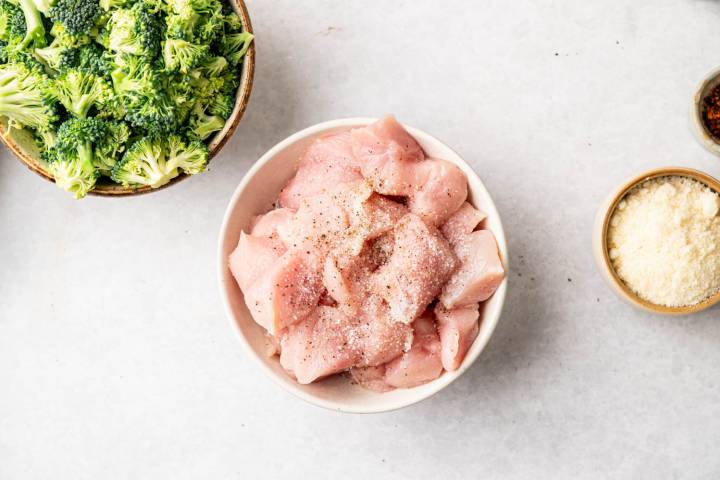 A bowl of raw, seasoned chicken pieces is centered, flanked by a bowl of fresh broccoli florets and a bowl of grated parmesan cheese on a light surface.