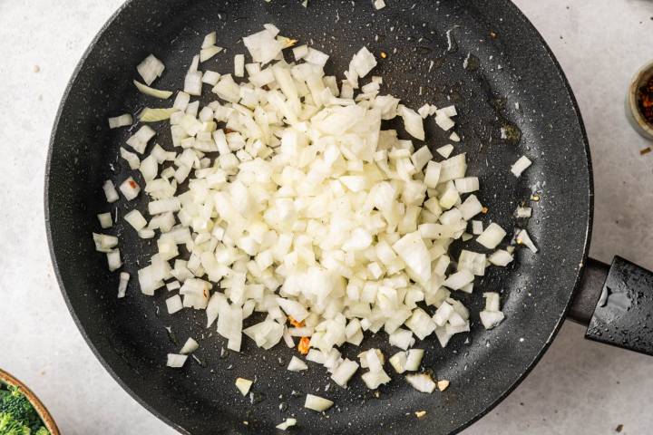 Chopped onions being sautéed in a black non-stick pan on a light countertop, with a small jar of spices partially visible in the corner.