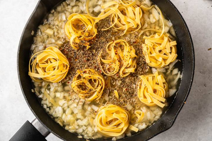 A black pan filled with rolled nests of uncooked tagliatelle pasta, surrounded by diced onions and sprinkled with mixed herbs and spices in broth.