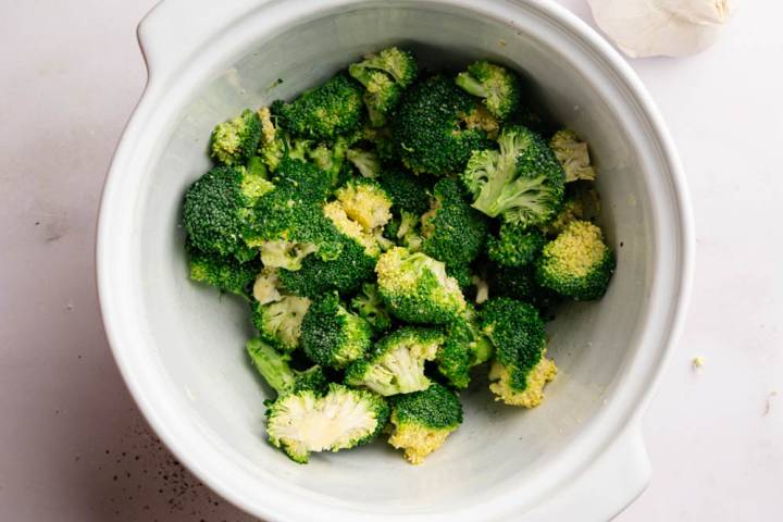 Fresh broccoli florets in a white bowl are surrounded by garlic, a wooden pepper grinder, a small dish of salt, and a bottle of olive oil.