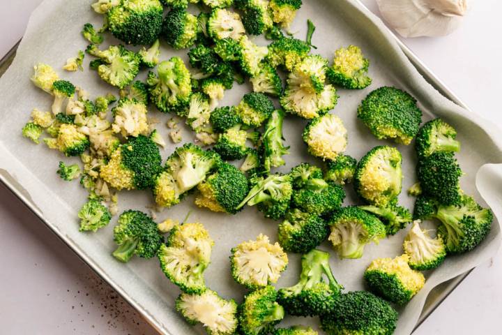 Broccoli florets are spread on a parchment-lined baking sheet, ready to roast. Nearby are a wooden pepper mill, salt dish, olive oil, and a garlic bulb.