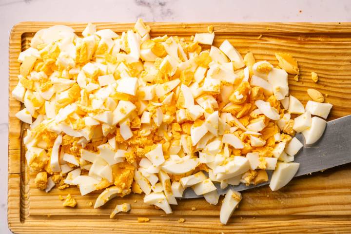 Chopped hard-boiled eggs scattered on a wooden cutting board with a visible knife. 