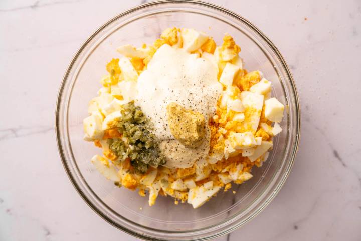 A glass bowl filled with chopped hard-boiled eggs, mayonnaise, mustard, relish, and pepper on a marble countertop, ready for mixing.