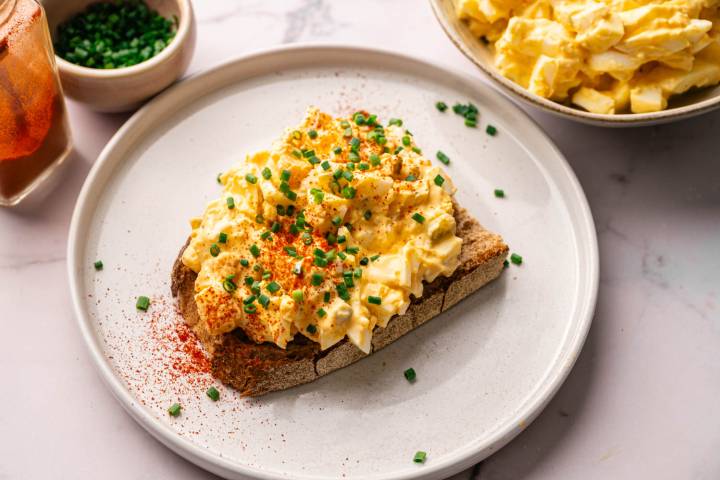 Egg salad on rye toast, garnished with chopped chives and paprika on a white plate. Nearby are bowls of more egg salad and chives.