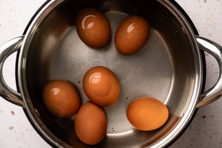 Six brown eggs are placed inside a stainless steel pot on a light speckled countertop, ready to be boiled.