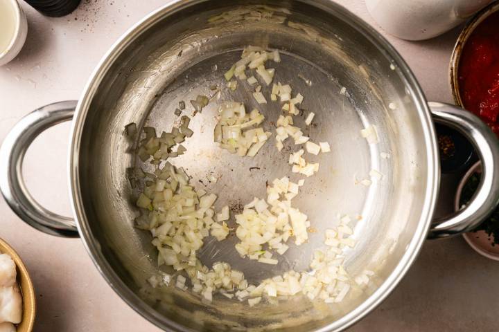 Chopped onions sautéing in a large stainless steel pot, surrounded by various ingredients on a kitchen counter, suggesting meal preparation.