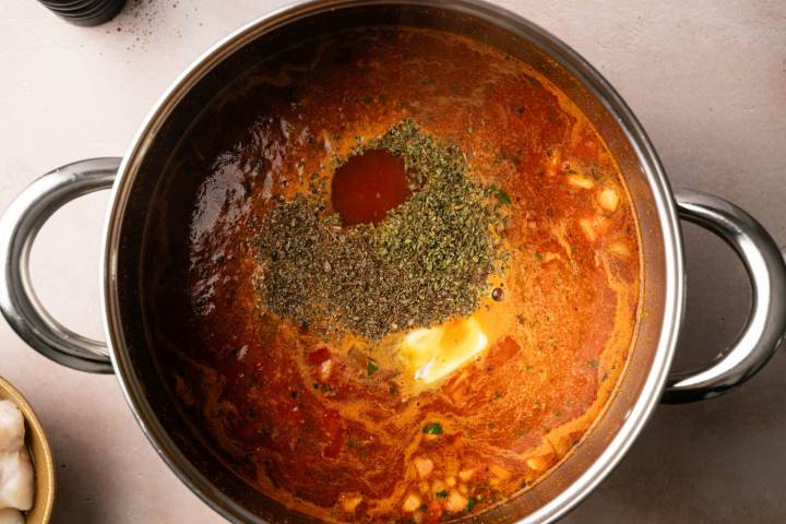 A pot filled with rich, red curry sauce topped with a pile of herbs, a dollop of butter, and a splash of hot sauce, suggesting a flavorful meal.